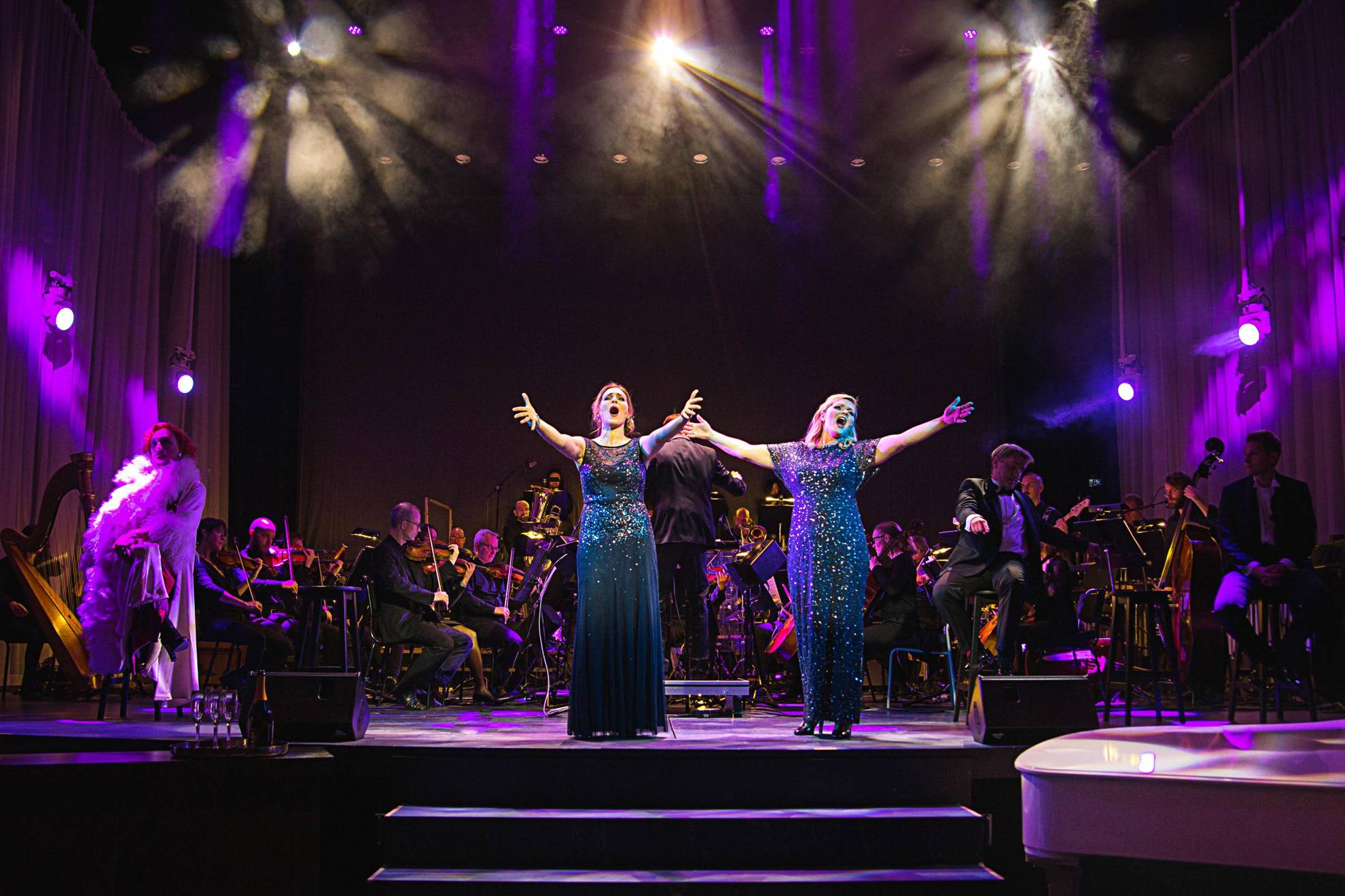 Twee vrouwen in blauwe glitterjurken zingen op een podium tijdens een optreden. Achter hen staat een orkest.