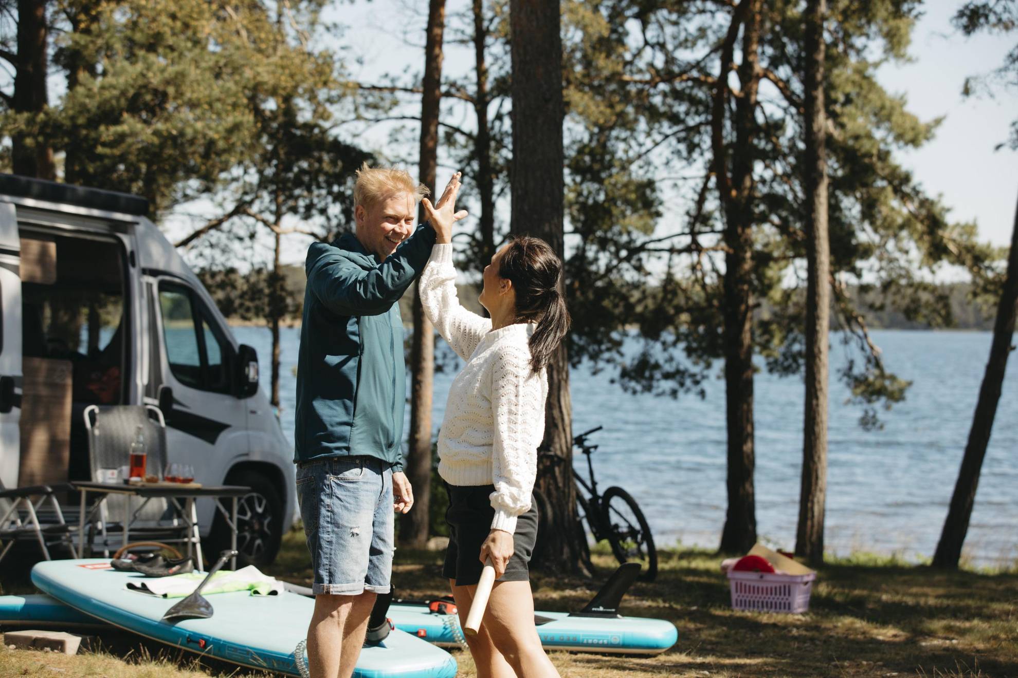 Een man en een vrouw high fives die in de zomer buiten hun camper staan. Er liggen op de grond staande paddleboards.