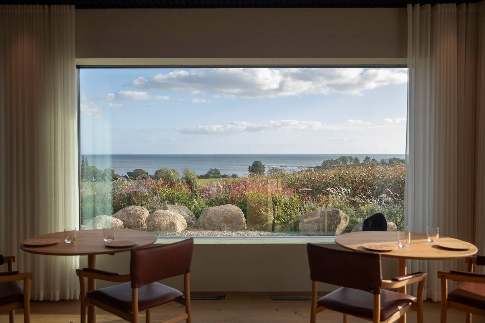 Twee tafels met stoelen in restaurant Vyn, staande voor een groot raam en uitzicht op de zee op een zomerdag.