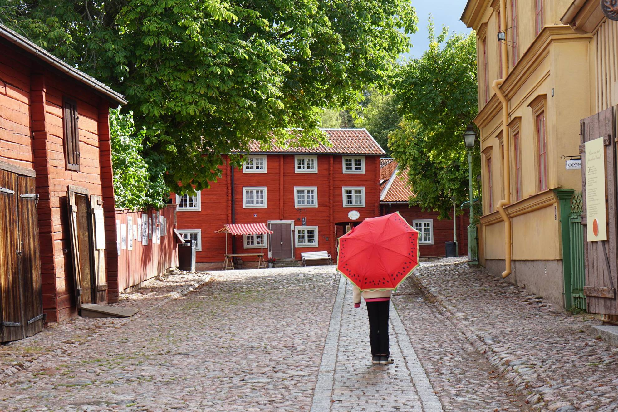Een persoon loopt met een rode paraplu op een geplaveid pad tussen oude huizen