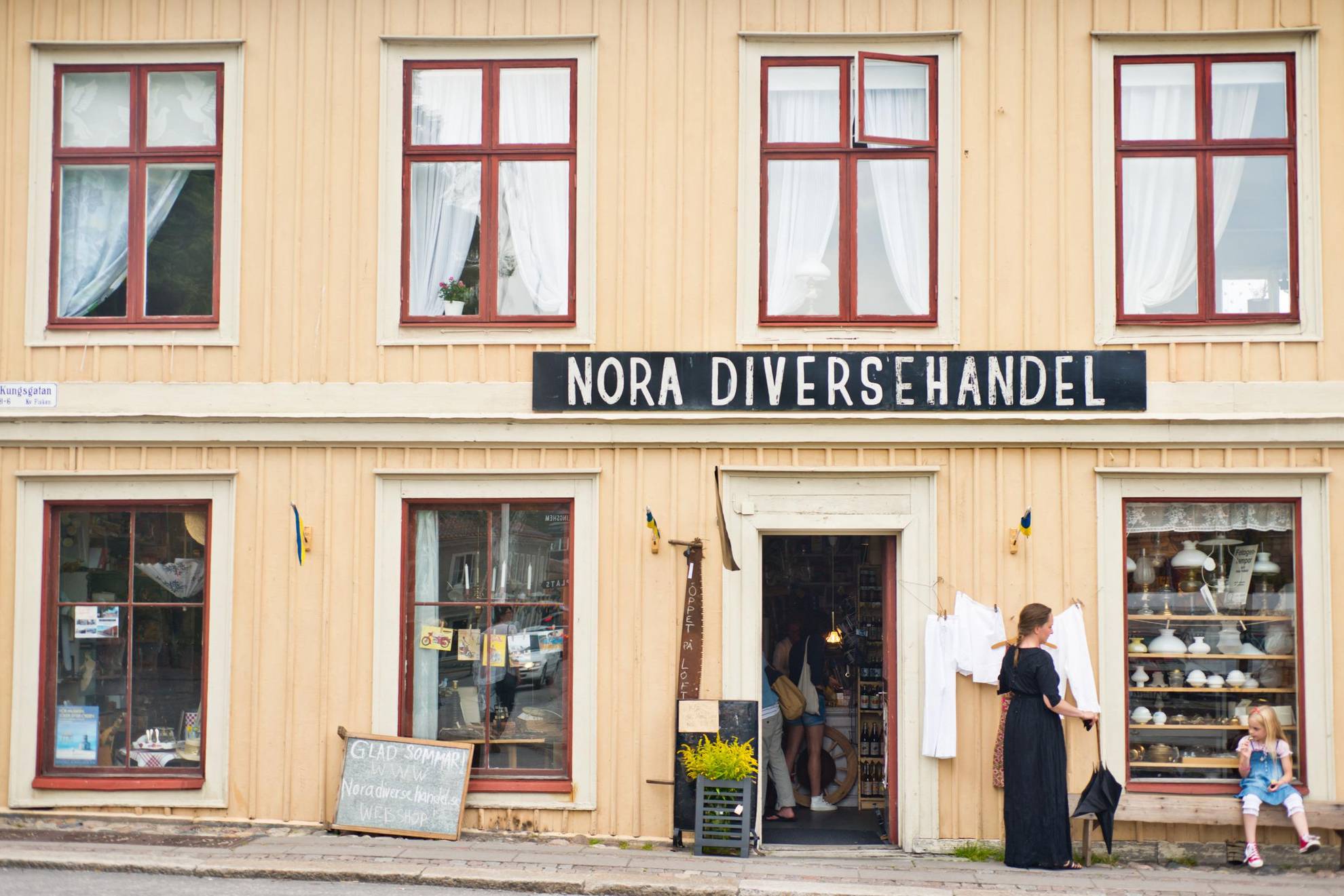 Een kleine winkel in een geel houten huis aan een straat in een oude stad. Op een bord boven de ingang staat Nora Diversehandel. Een vrouw staat en een meisje zit op een bankje voor het huis.