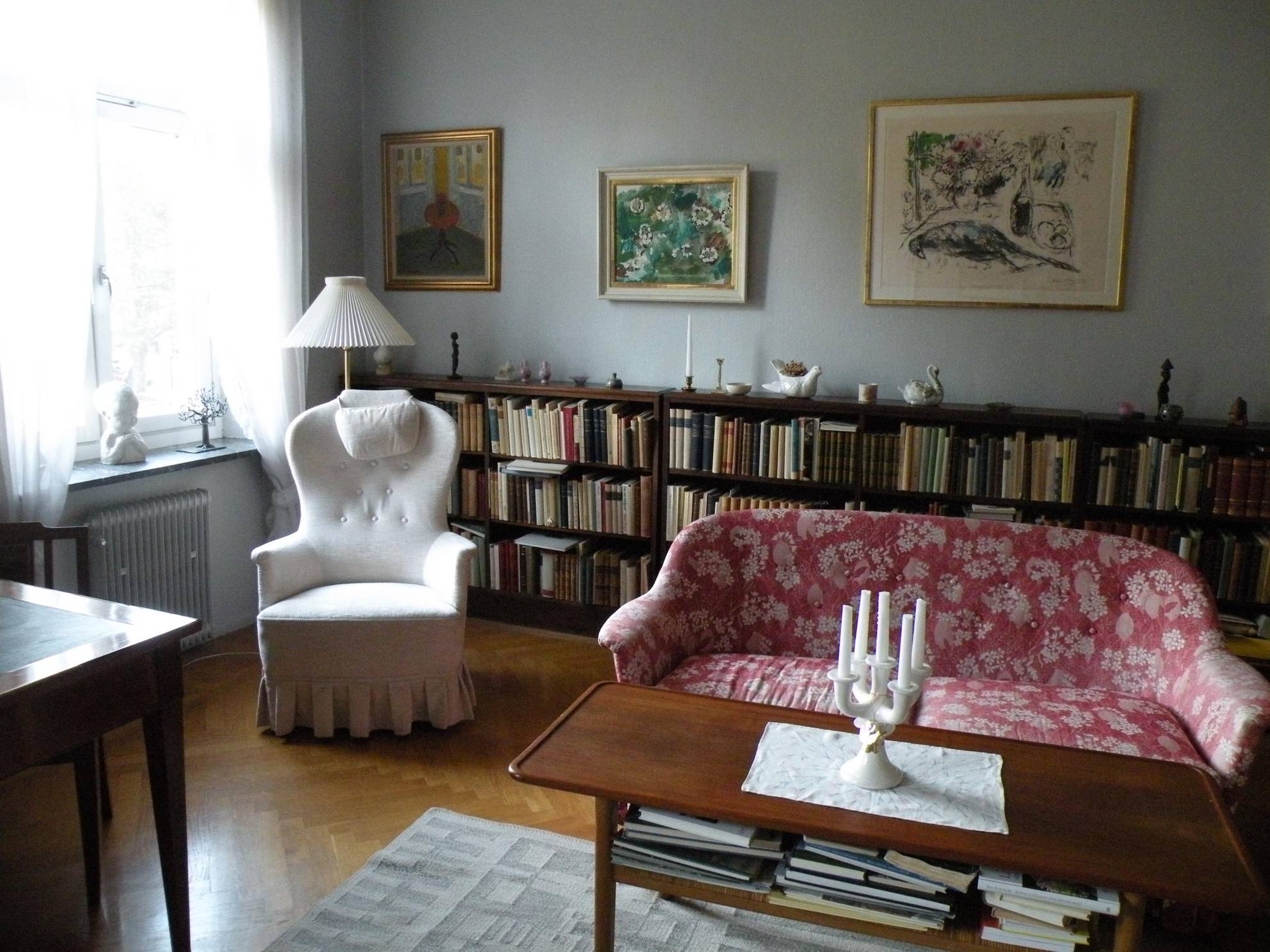 De woonkamer van Astrid Lindgren, met een leesstoel, een grote boekenplank met bibelots erop, een rode bank met patroon en een paar schilderijen aan de muur.