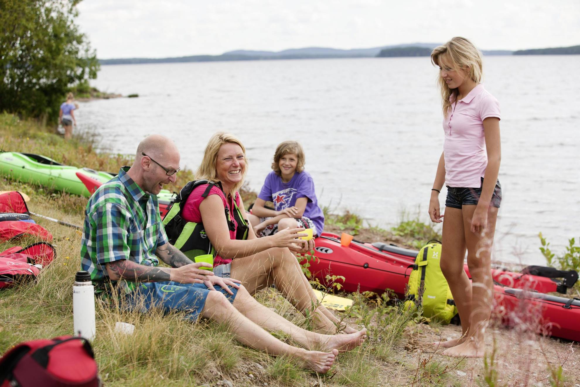 Een man, een vrouw, een jong meisje en een jongen rusten aan de oever van een meer naast hun kajaks.