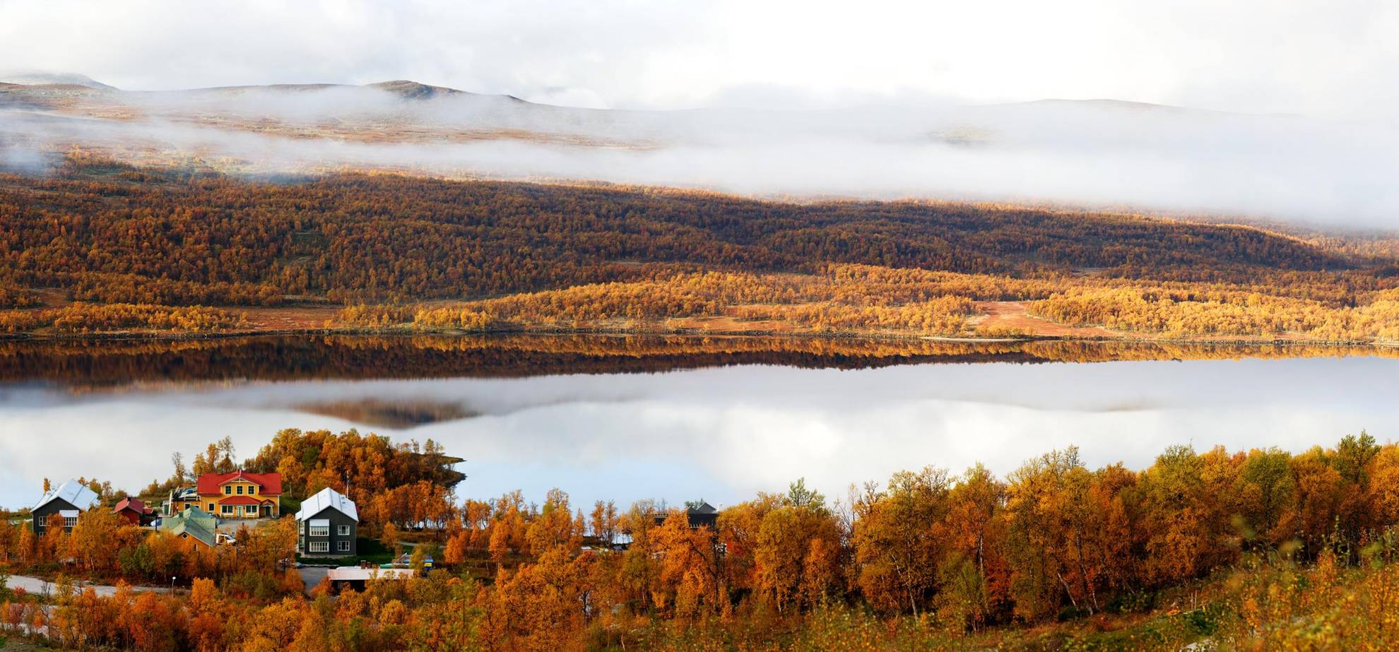 Een prachtig herfstbeeld van een hotel aan een meer in de bergen. De bomen zijn oranje en er hangt mist over de berg aan de andere kant van het meer.