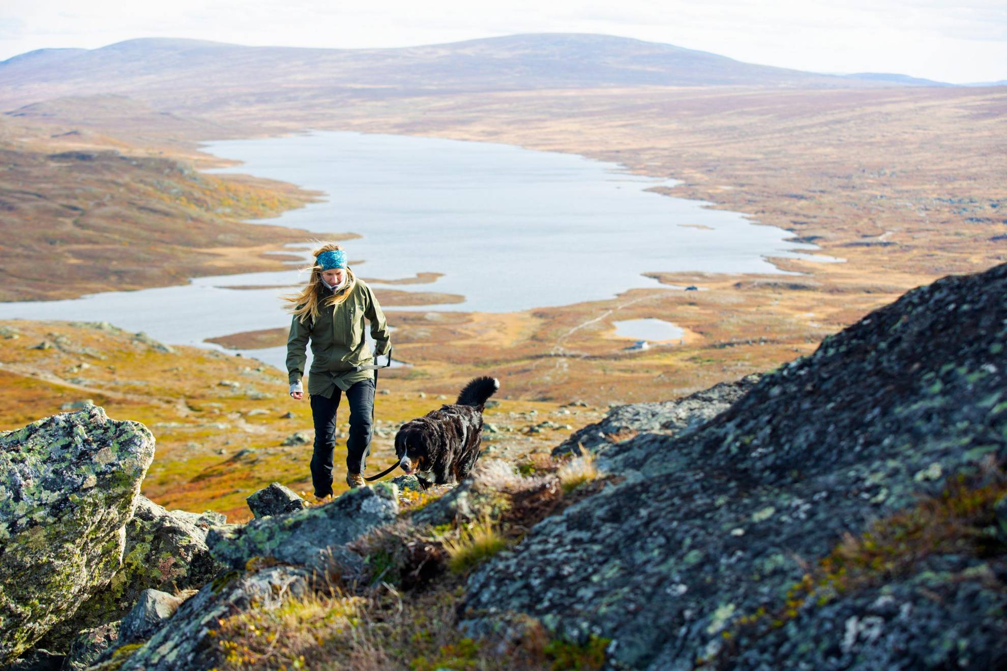 Een vrouw die met een hond in de bergen wandelt. Uitzicht op bergen en een meer op de achtergrond.