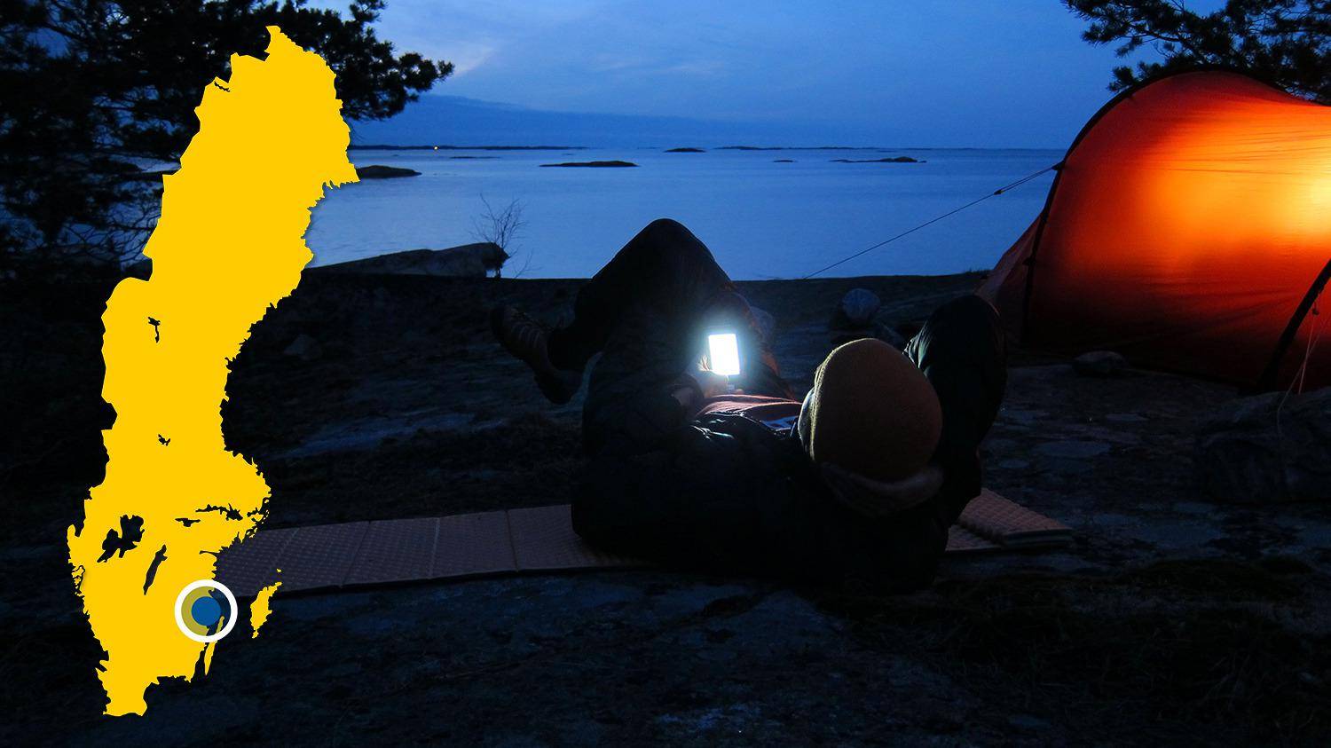 Op een strand aan het water staat een tent opgezet. Het is nacht en een persoon ligt naast de tent naar zijn telefoon te kijken.