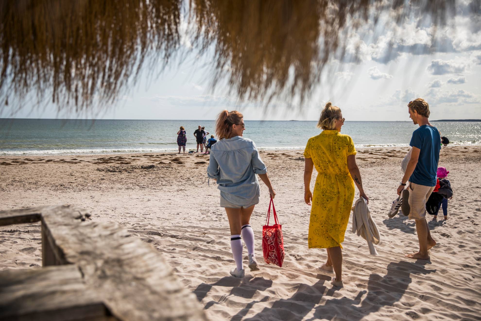 Twee vrouwen en een man op weg naar het strand. Je ziet de zee en enkele mensen op het strand op de achtergrond.