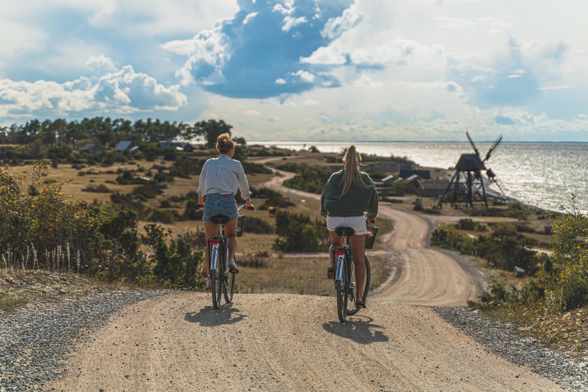 Twee vrouwen fietsen bij zonnig weer langs de kust van Öland met een windmolen op de achtergrond