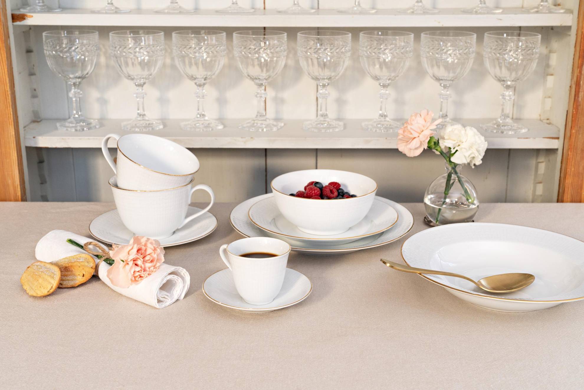 Een serviesservice genaamd "Swedish Grace" op een tafel. Borden in verschillende maten een kom met bessen, twee theekopjes en een kopje koffie. Daarachter is een witte plank met verschillende wijnglazen.