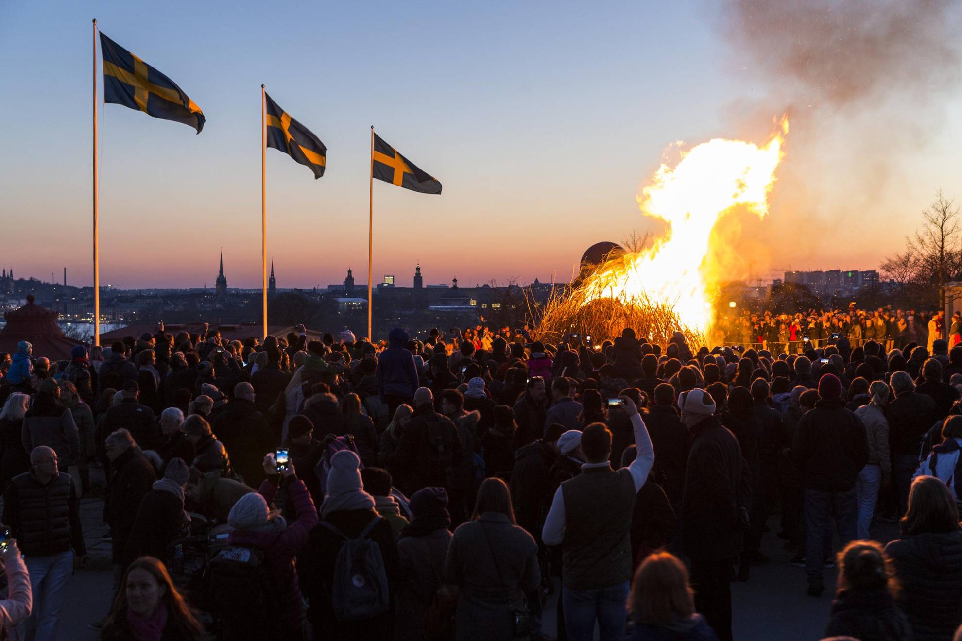 Mensen hebben zich verzameld rond een vreugdevuur om Walpurgisnacht te vieren. Drie Zweedse vlaggen wapperen in de wind.