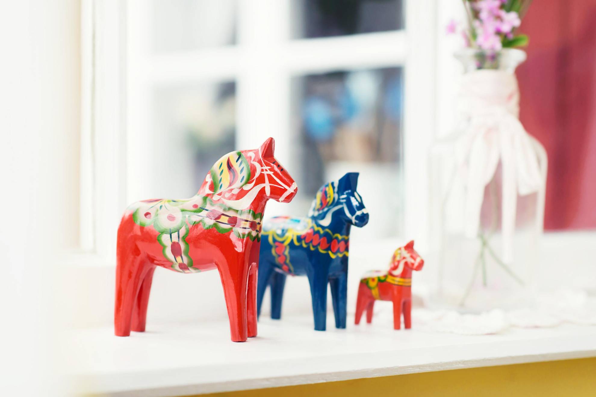 Drie Dala-paarden in verschillende maten op een vensterbank. De grootste en de kleinste zijn in rood geschilderd en de middelste in blauw met kubit-versieringen.