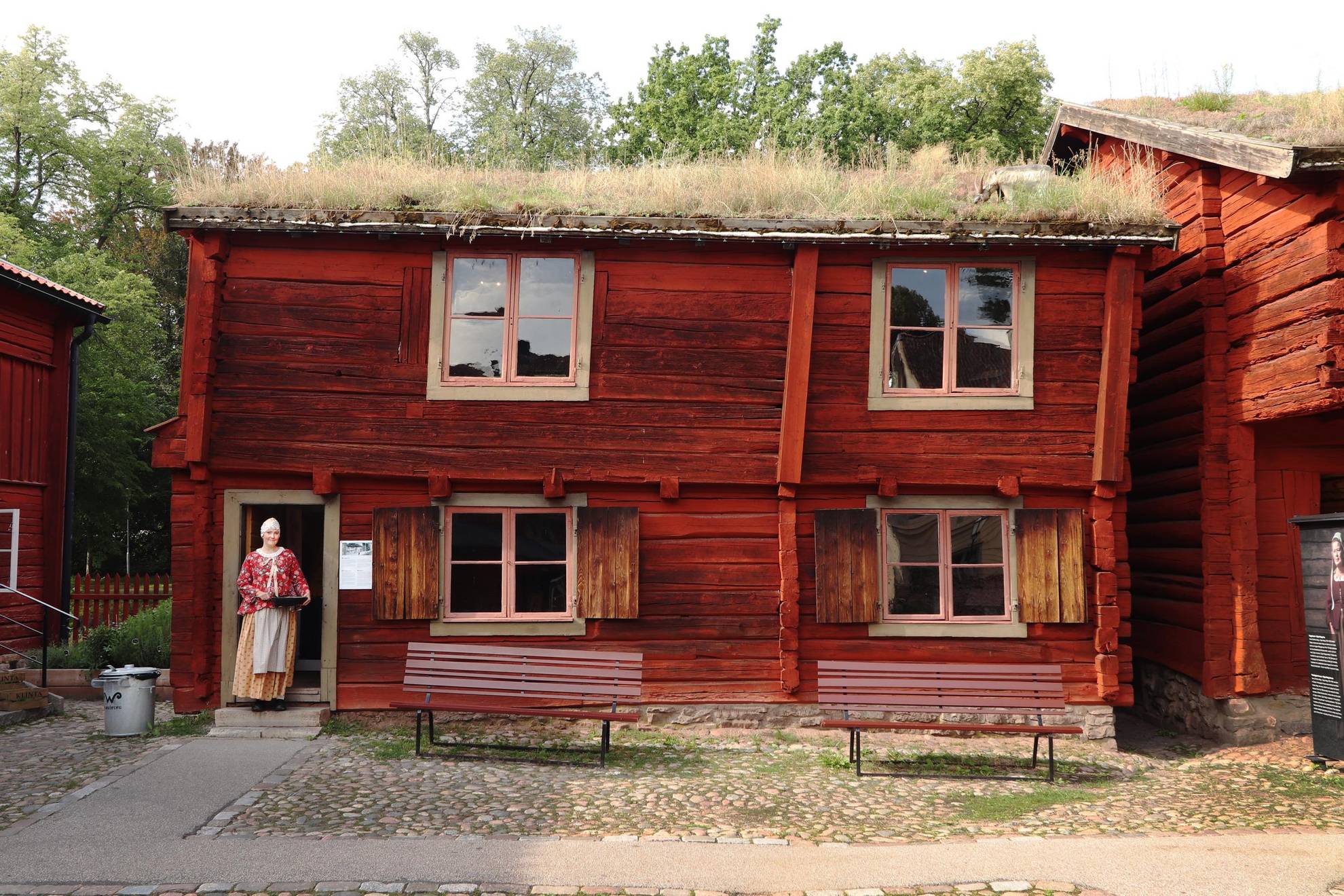 Een oud rood geschilderd houten huis met een zodendak. Bij de ingang van het huis staat een vrouw.