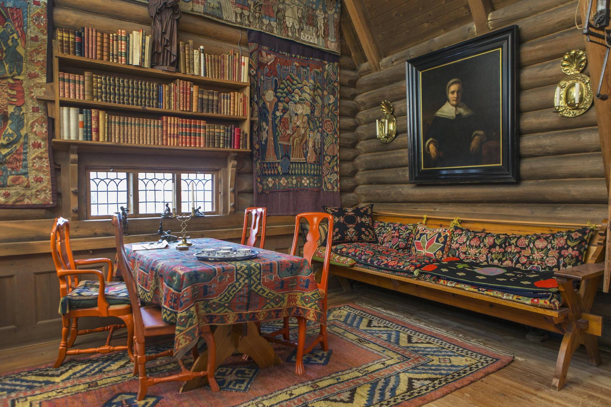 De kleurrijke eetzaal van Anders Zorn, met geweven wandtapijten, knalrode stoelen rond een tafel met een geweven doek, een boekenplank en een houten bank.
