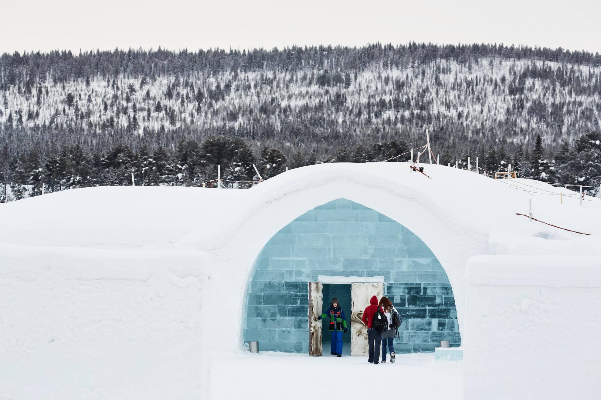 In een boslandschap betreden mensen het IJshotel, een gebouw gemaakt van ijs en opeengepakte sneeuw, via een deur gemaakt van ijs, rendierhuid en gewei.