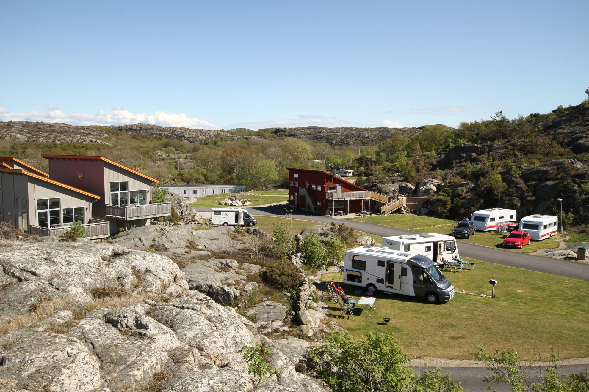 Campers geparkeerd op een camping naast een stenen klif. Op de achtergrond zijn drie grote kampeerhutten met schuine daken te zien.