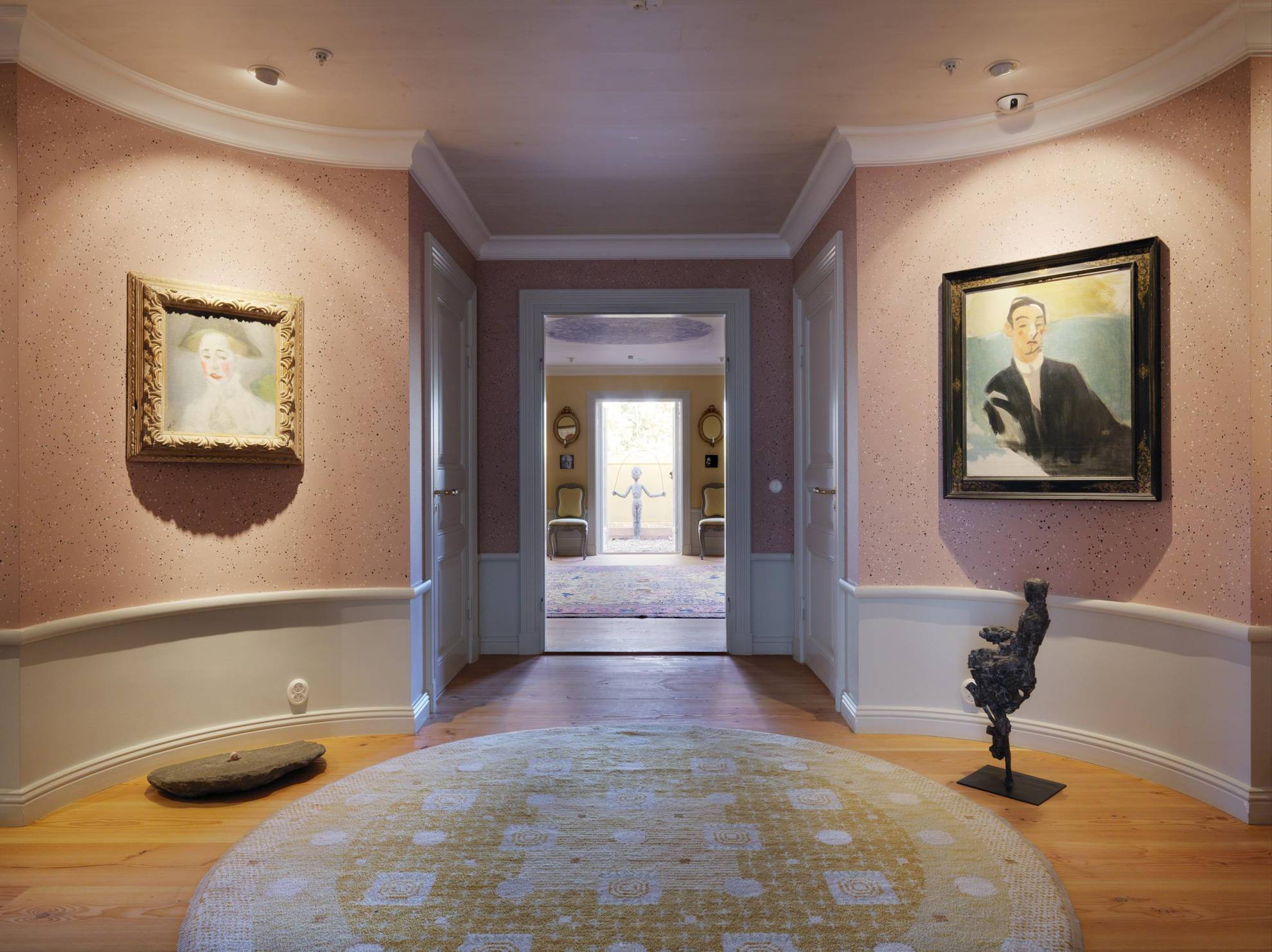 Een gang met roze wandtapijten en schilderijen aan weerszijden van een deuropening die naar een andere kamer leidt.