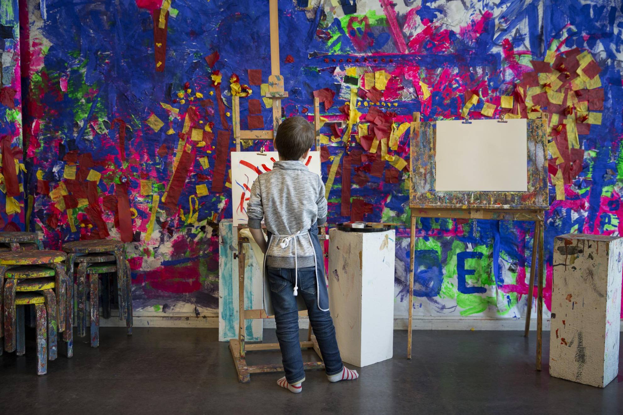 In Room for Children is een jongen aan het schilderen op een doek dat op een ezel is opgesteld. De achtergrondmuren en stoelen zijn bedekt met verf.