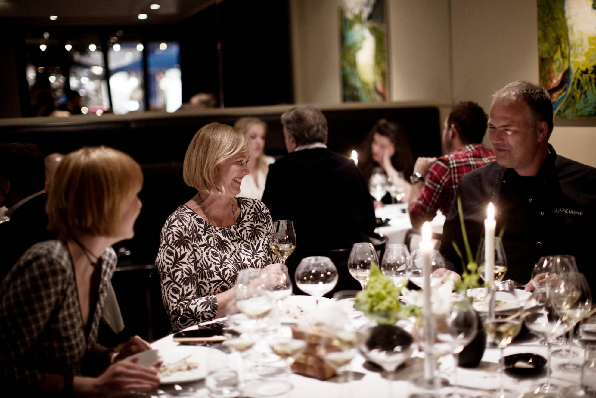 Twee vrouwen en een man dineren in een restaurant. Je ziet ook enkele gasten aan een tafel achteraan.