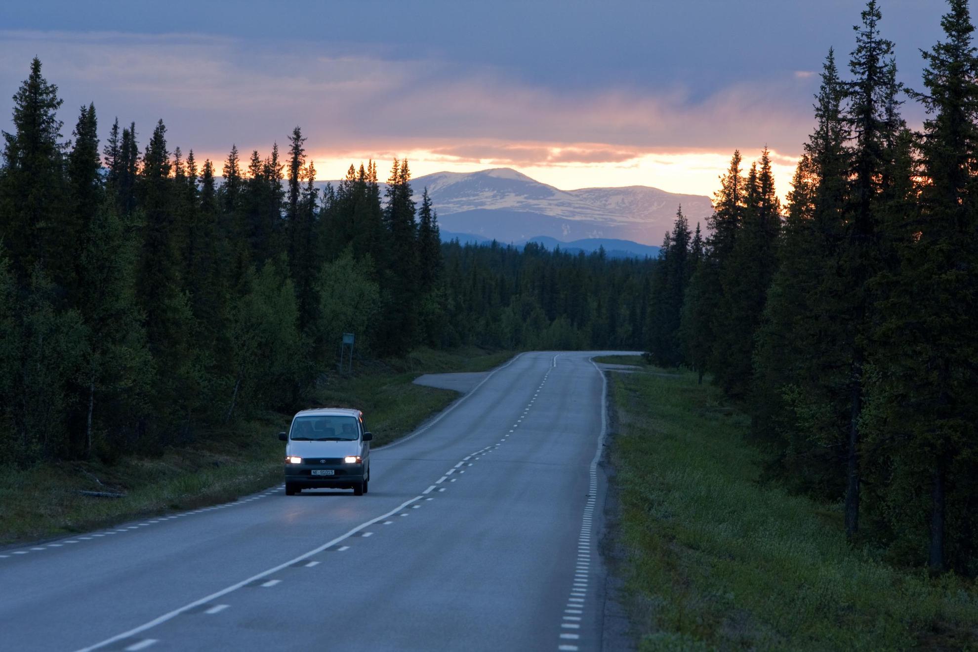 Een minivan rijdt langs een weg tijdens de middernachtzon in Noord-Zweden, bossen aan beide kanten en bergen op de achtergrond.