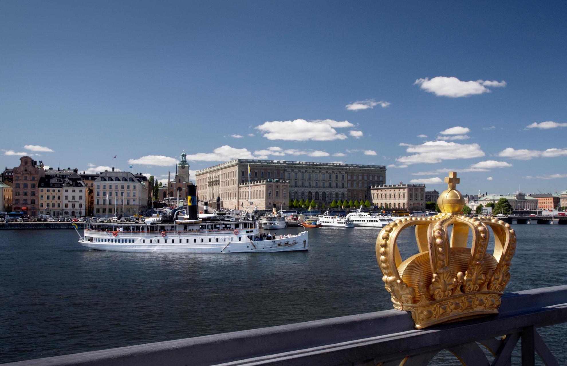 Een gouden kroon op de omheining van de Skeppsholmenbrug met het Koninklijk Kasteel en veerboten op de achtergrond.