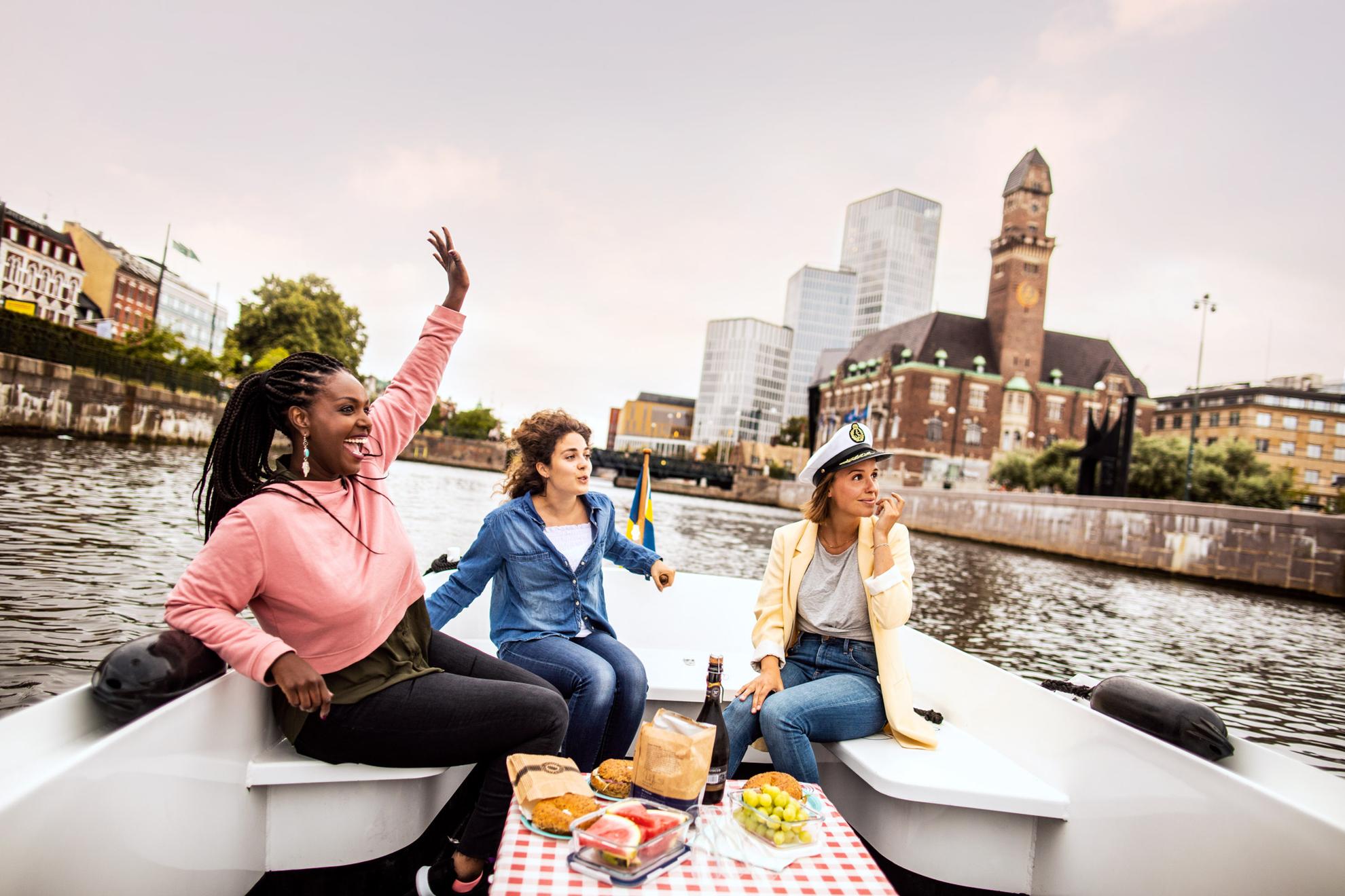 Drie jonge, gelukkige vrouwen op een excursie in een boot op het kanaal van Malmö met Malmö Live en het centraal station op de achtergrond.