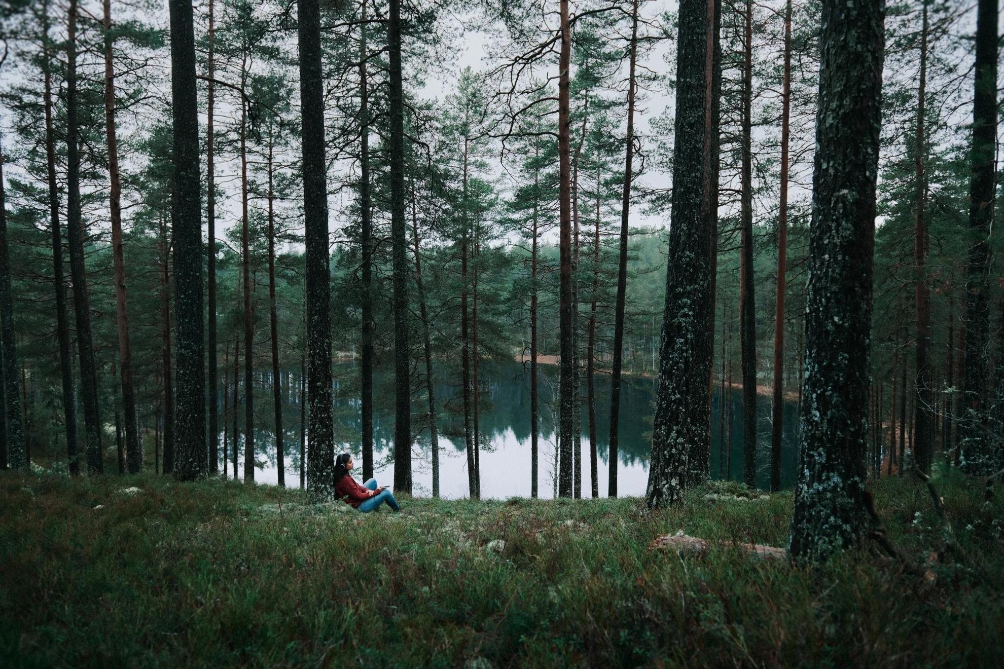 Een vrouw die een koptelefoon draagt, rust tegen een dennenboom naast een meer. Het weer is somber.