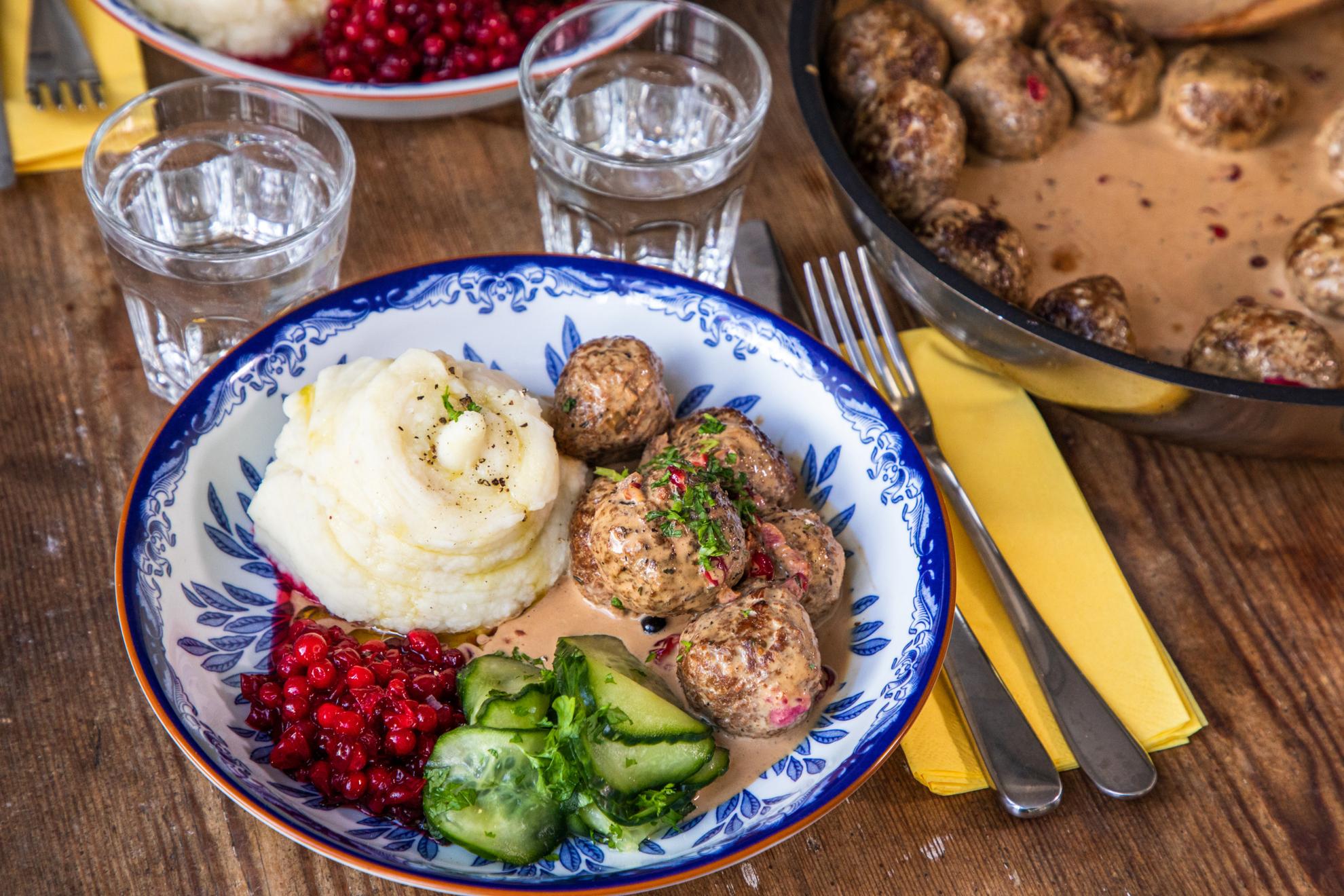Een bord met vegan gehaktballetjes, saus, ingelegde komkommer, rode bosbessenjam en aardappelpuree.