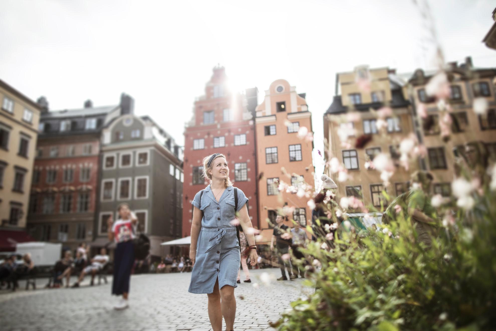 Een vrouw loopt op een plein in de Oude Stad. Op de achtergrond zie je oude stenen huizen en vooraan staat een bloeiende plant.