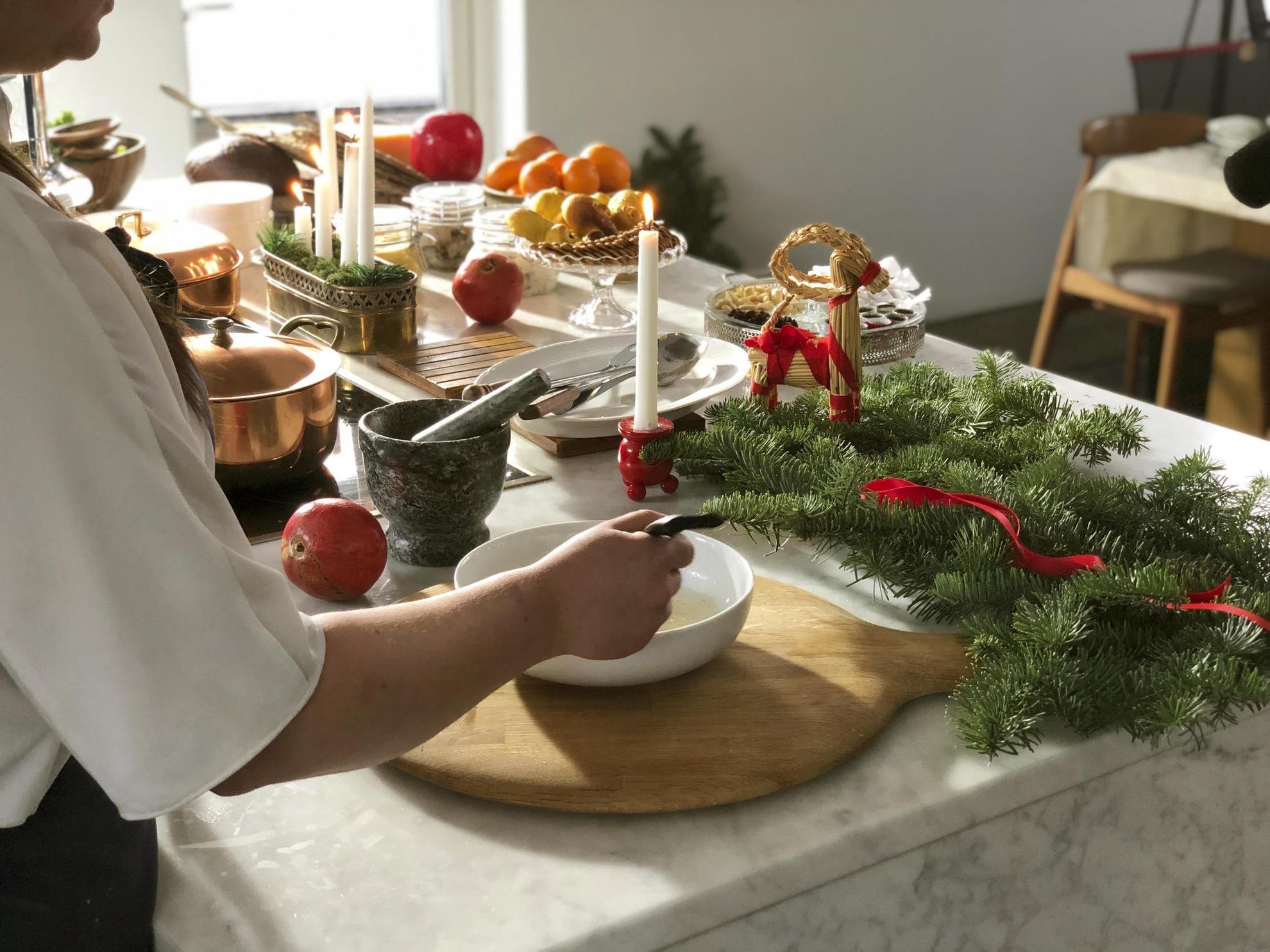Een vrouw die een kerstlunch klaarmaakt. Op de tafel voor haar staan diverse schalen, koperen potten, adventskandelaar, brandende kaarsen, dennentakjes en een klein kerstgeitje.