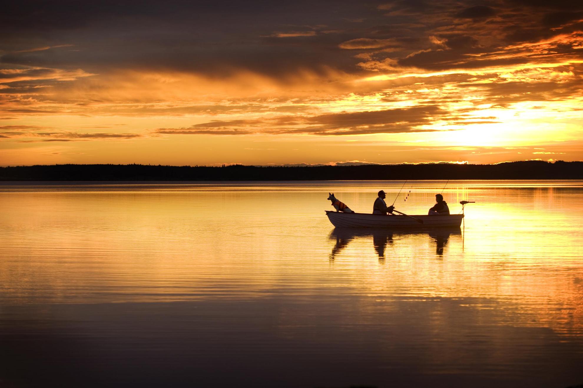 Twee mannen en een hond in een kleine boot op een meer vissen in de middernachtzon. De oranje lucht weerspiegelt in het meer.
