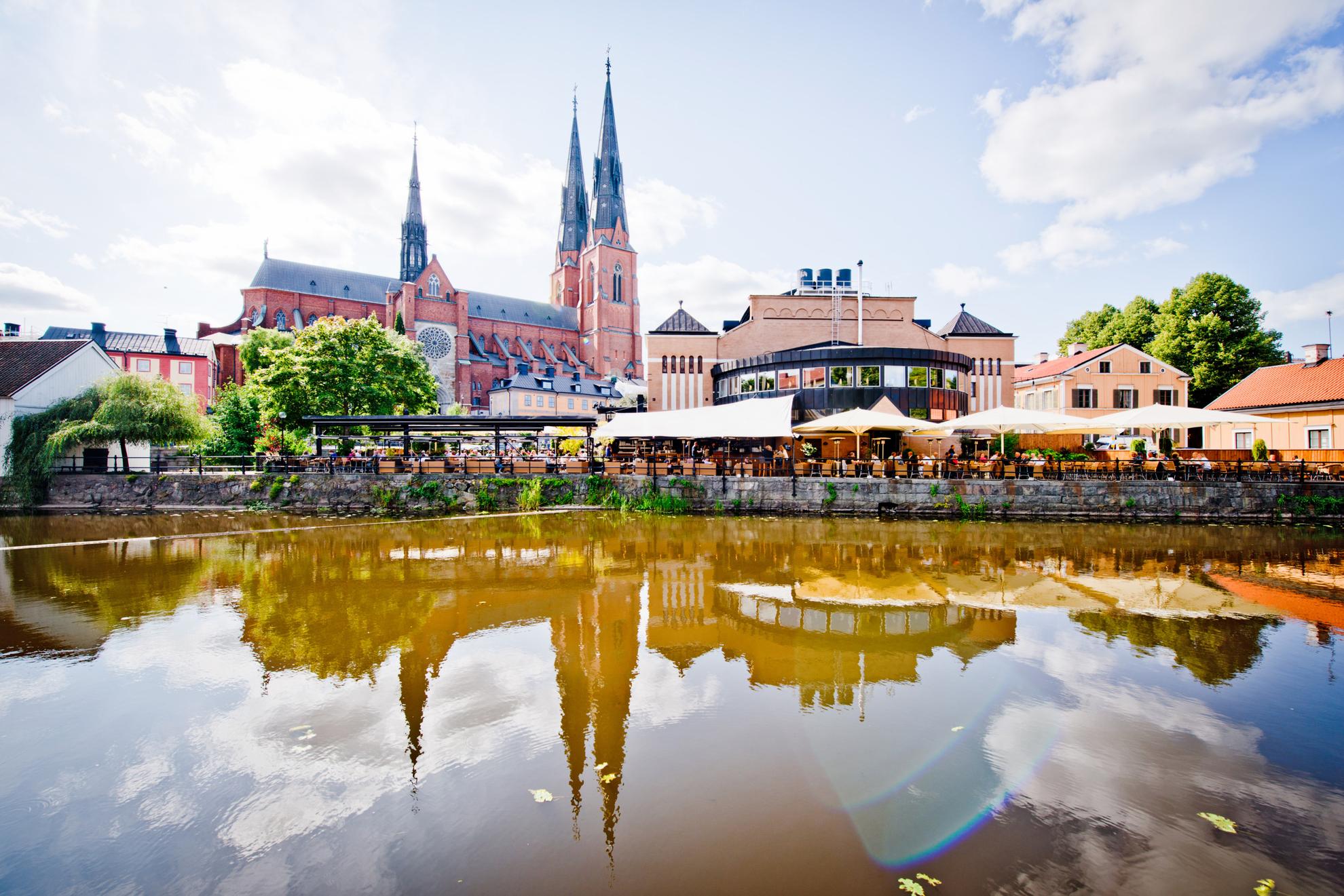Op de achtergrond rijst de kathedraal van Uppsala de lucht in. Ervoor staan ​​andere gebouwen en een buitenterras met veel stoelen. De gebouwen van de stad worden weerspiegeld in het water van de rivier de Fyris.