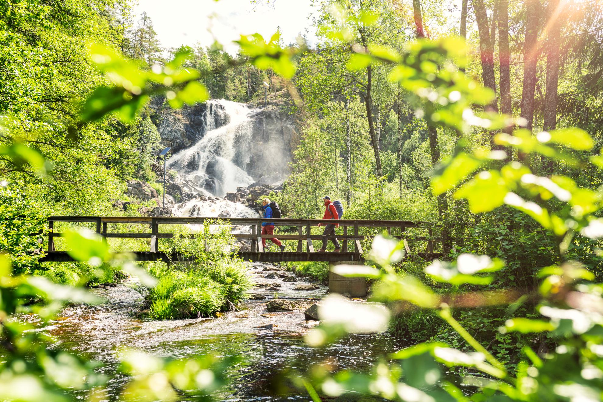 Twee mensen lopen over een houten brug in een bos. Er is een waterval op de achtergrond.
