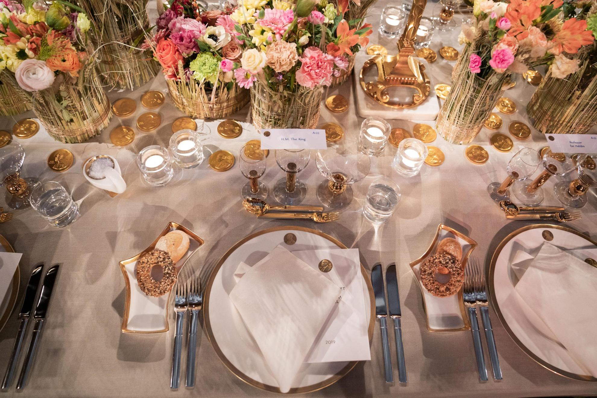 Op een tafel met een wit tafelkleed staat een wit servies met gouden details. In het midden van de tafel staan kleurrijke bloemen in vazen.