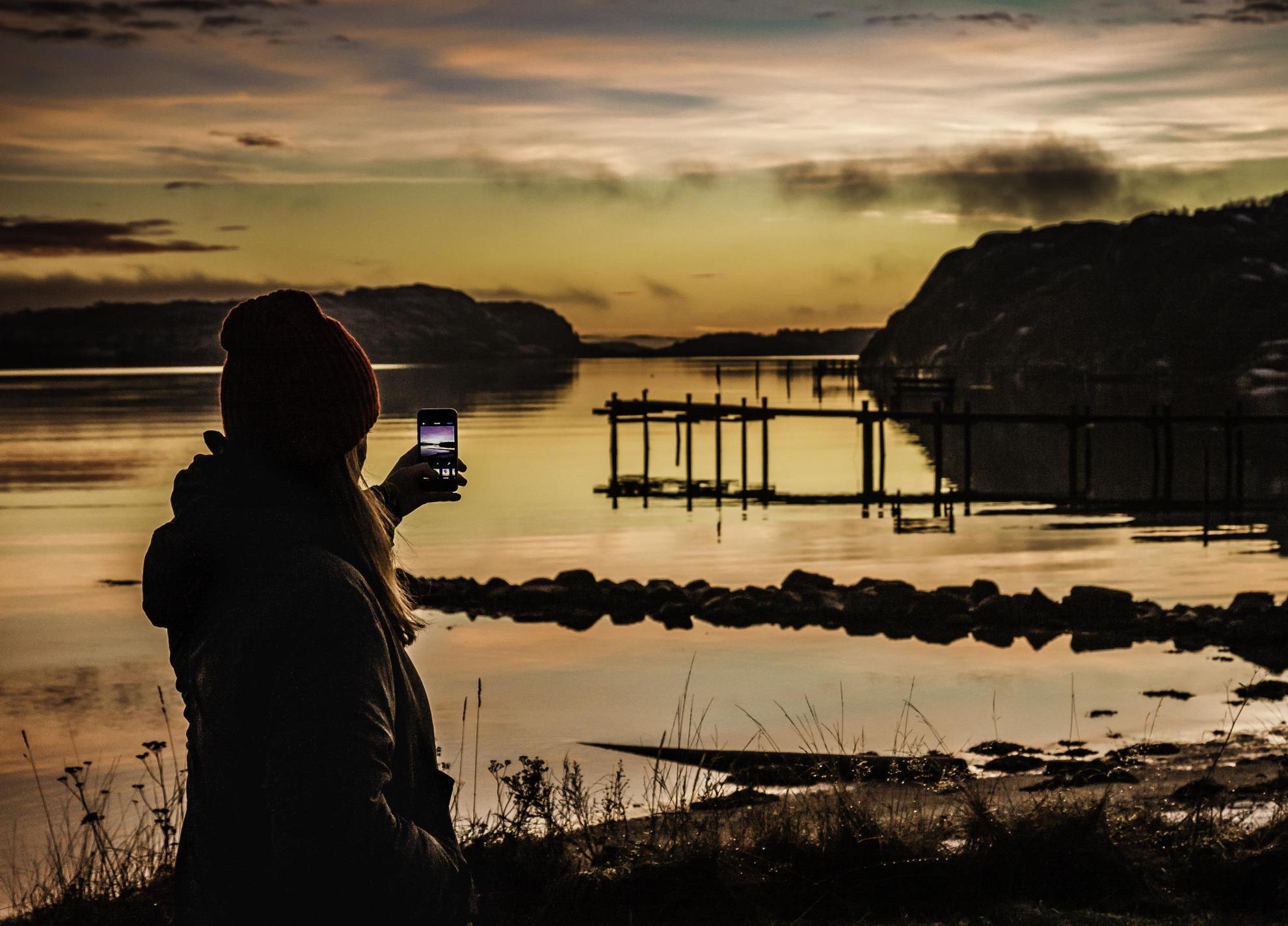 Een vrouw houdt een smartphone omhoog naar een zonsondergang aan het water aan de westkust van Zweden.
