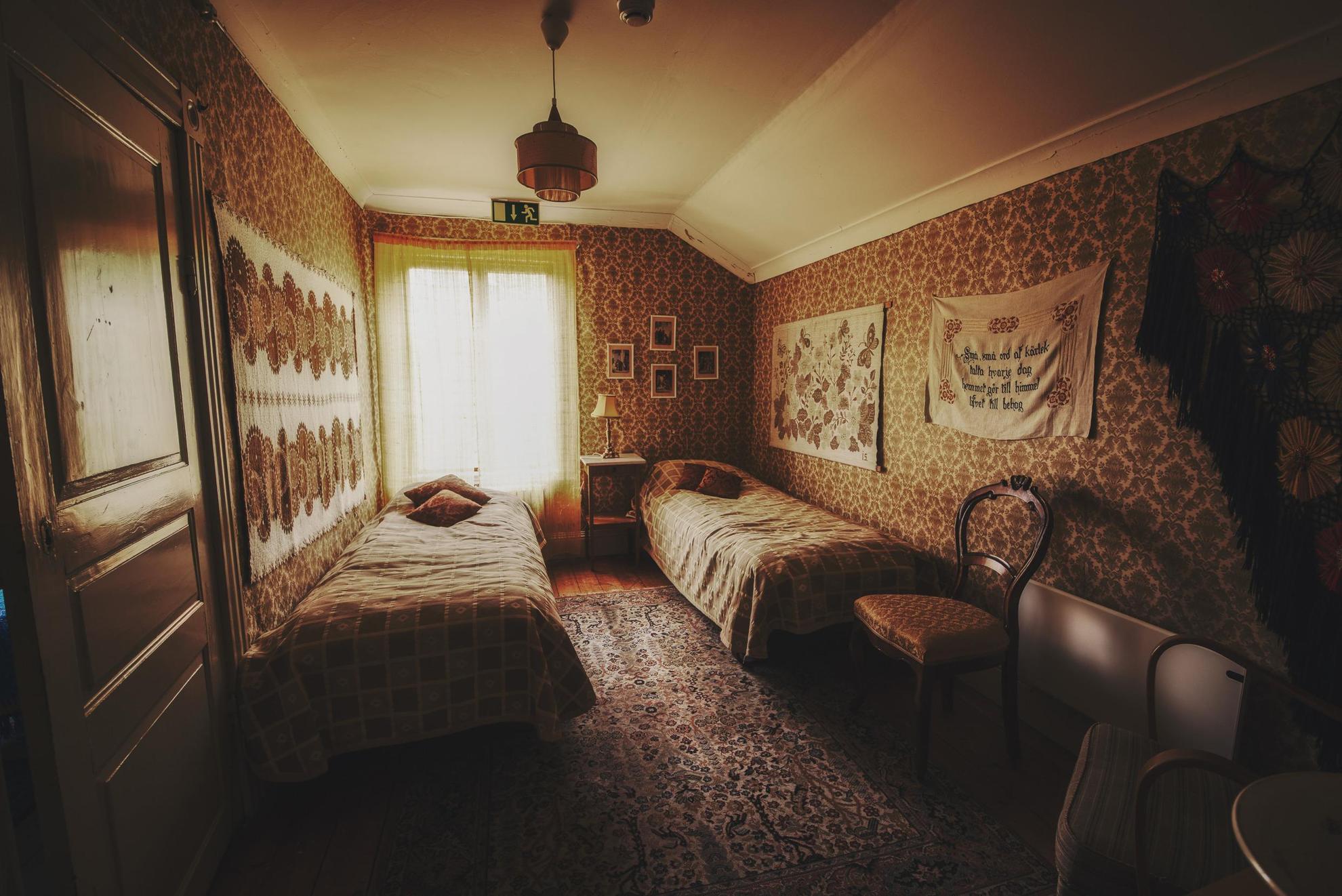 Een kamer met decoratief behang, schilderijen, een vloerkleed, een stoel en twee eenpersoonsbedden.