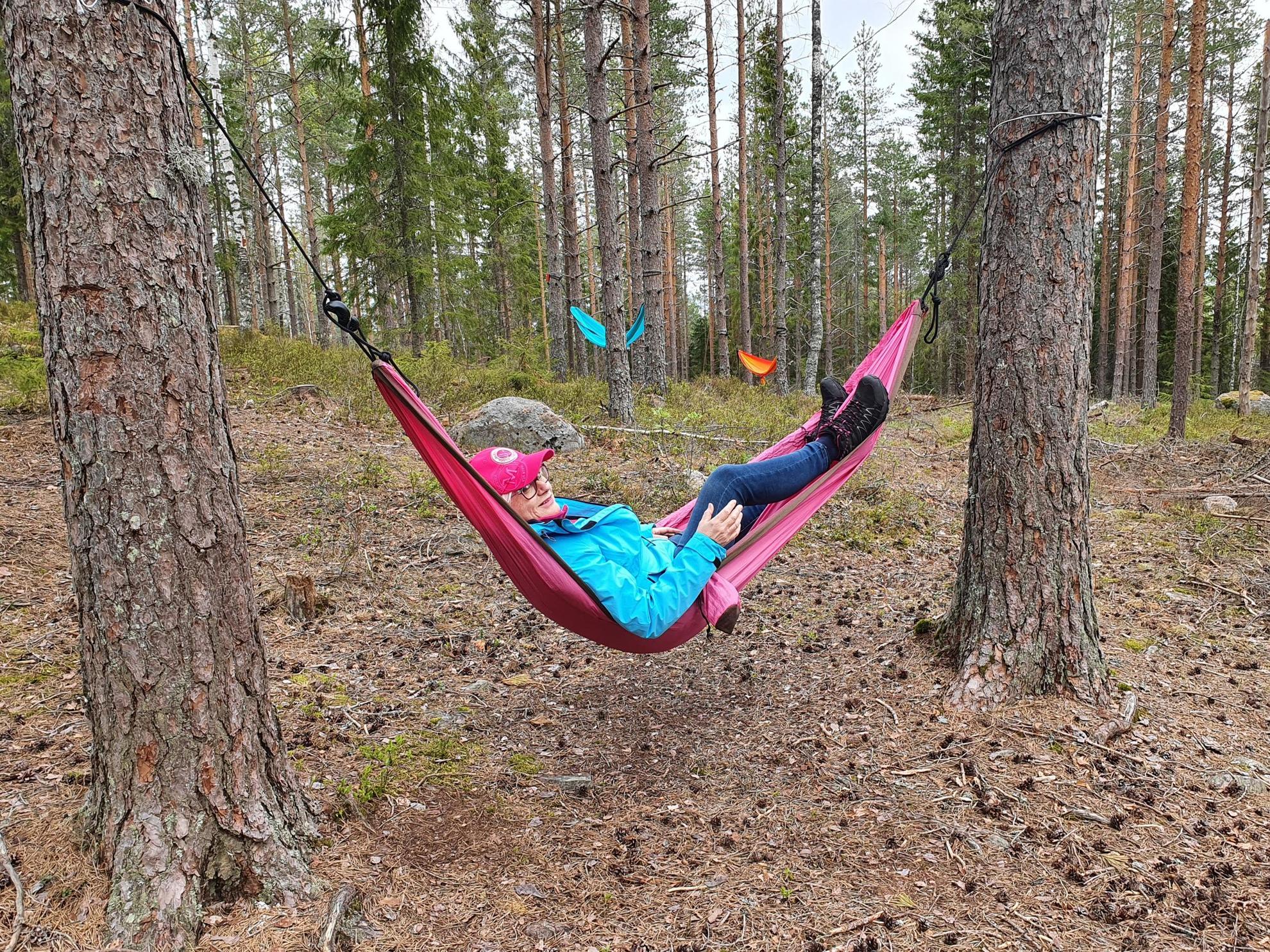 Een vrouw ontspant in een roze hangmat tussen twee dennen. Er is een blauwe en een oranje hangmat tussen de bomen op de achtergrond.