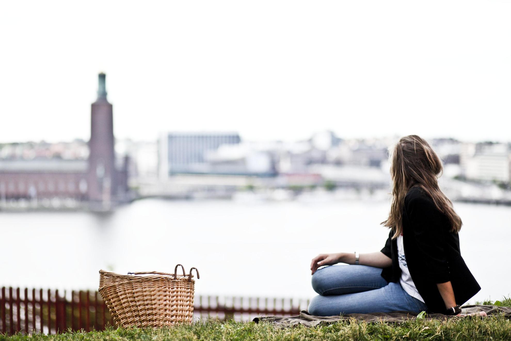 Een vrouw met een geweven mand zit in een park met uitzicht op het Mälarmeer en het stadhuis van Stockholm.