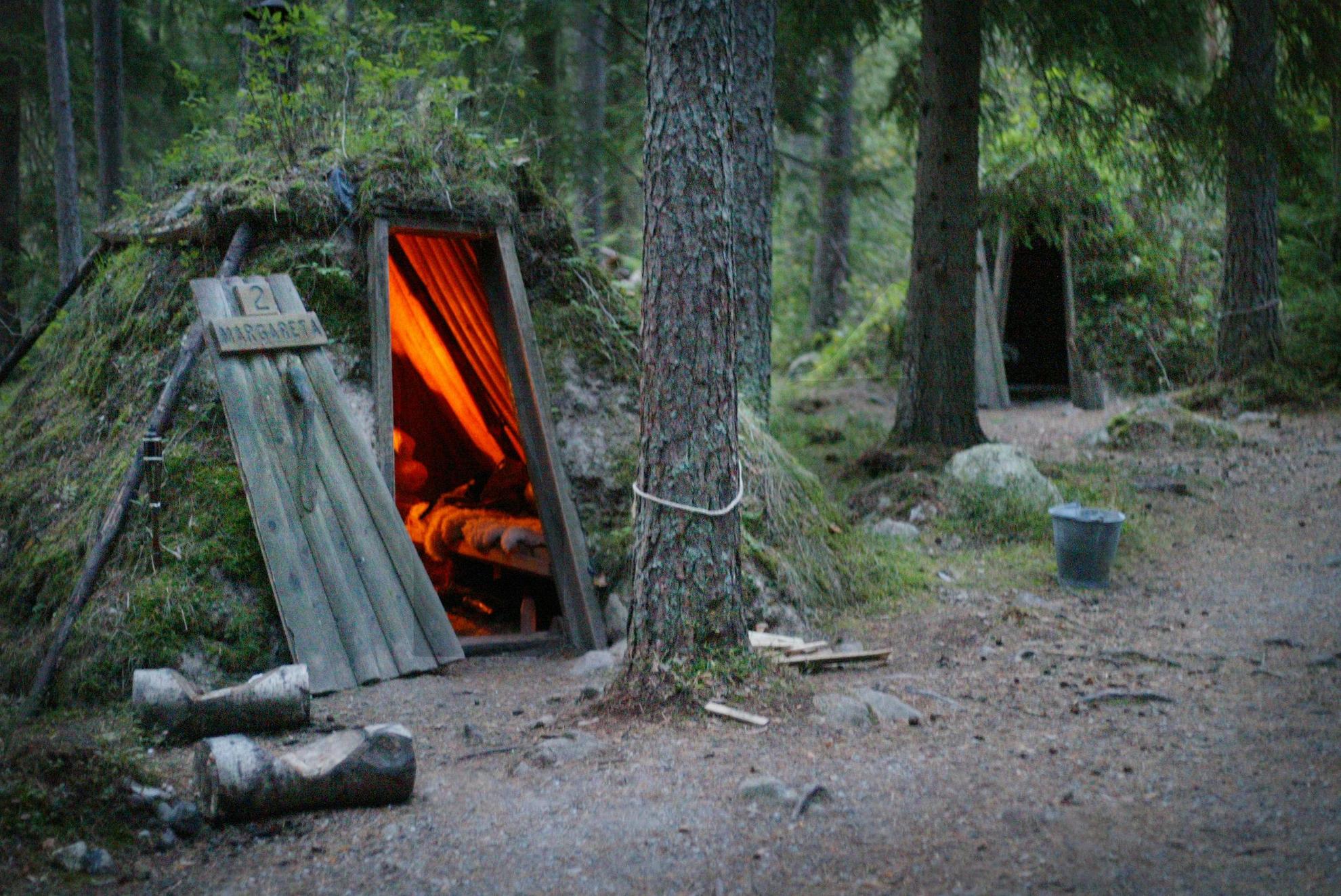 Twee boshutten bedekt met mos gelegen in een bos. In een van de hutten brandt een open haard.