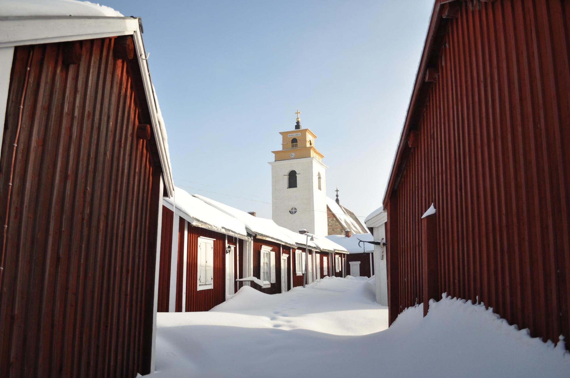Verschillende kleine houten huisjes leiden naar een witte stenen kerk. Sneeuw op de grond en huizen.