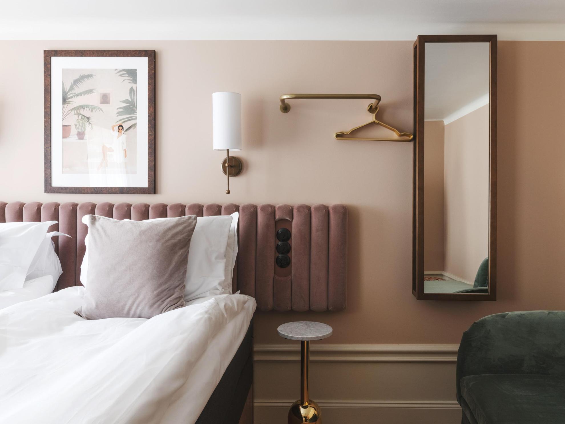 Een hotelkamer met beige geschilderde muren en een wit plafond. Een koperen wandlamp, een schilderij, een spiegel en een kleerhanger sieren de muren naast een tweepersoonsbed.