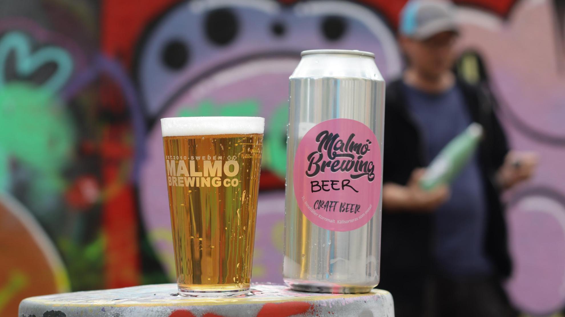 Een bierblikje en een glas gevuld met bier. Onscherp op de achtergrond is een persoon en een graffitimuur.