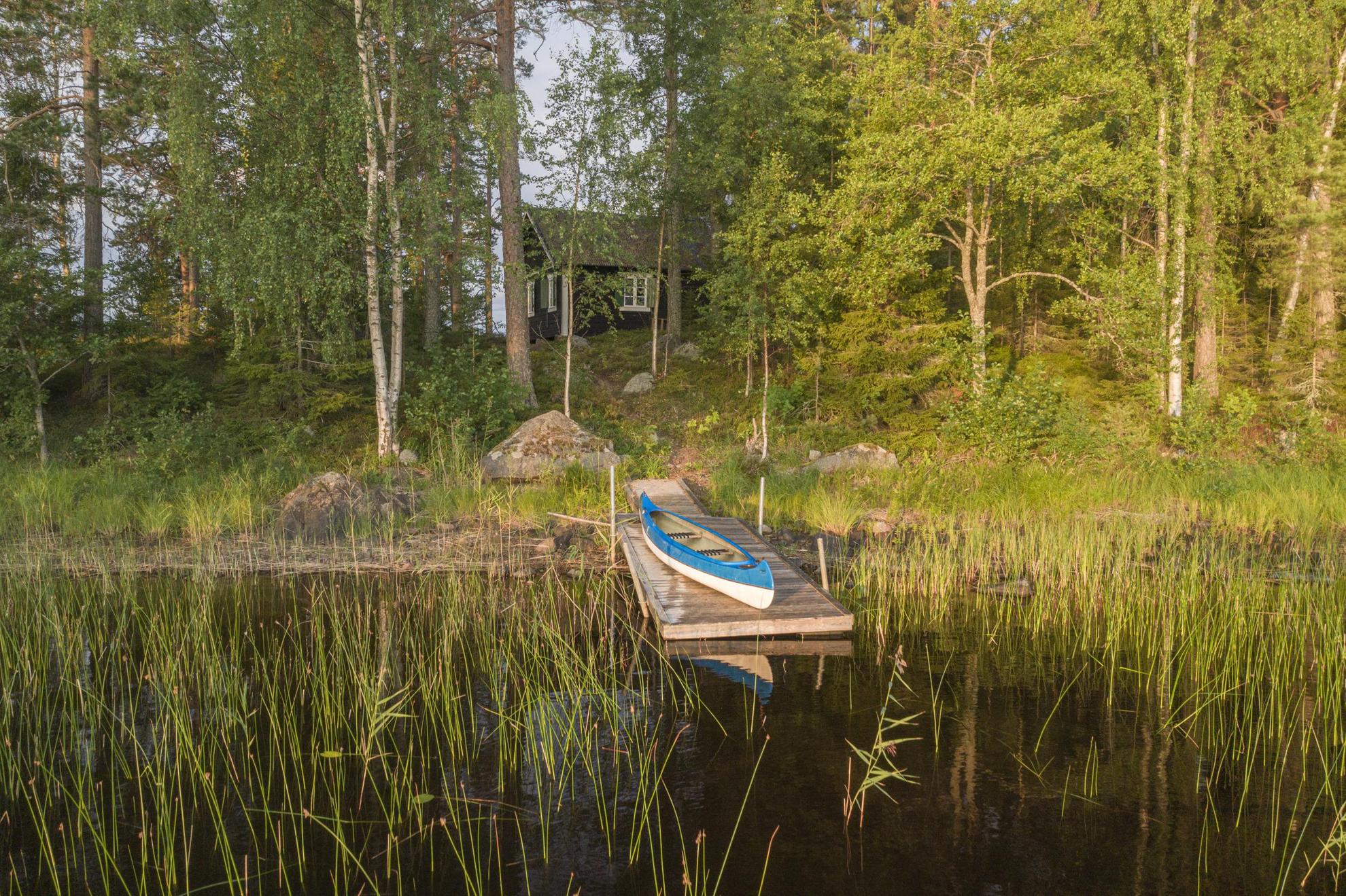 Een kano op een kleine steiger in het riet van een ondiep meer, waar je een houten hut kunt zien verborgen in het bos erachter.