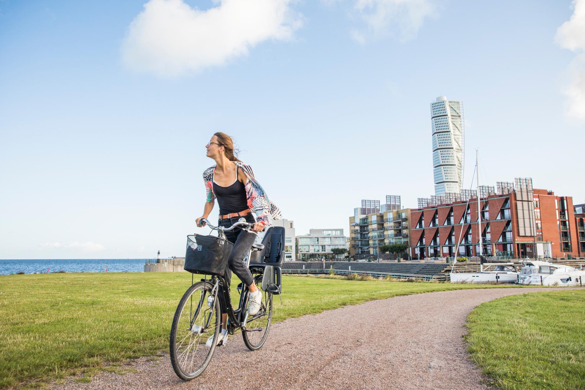 Een vrouw fietst op een loopbrug, hierachter zie je een woonwijk aan zee en in het midden een wolkenkrabber.