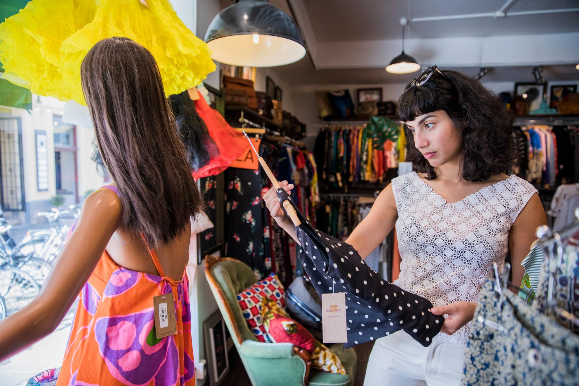 Een vrouw kijkt naar een blouse in een vintage winkel. In het raam naast haar staat een paspop en achter haar zie je heel veel verschillende kleding.