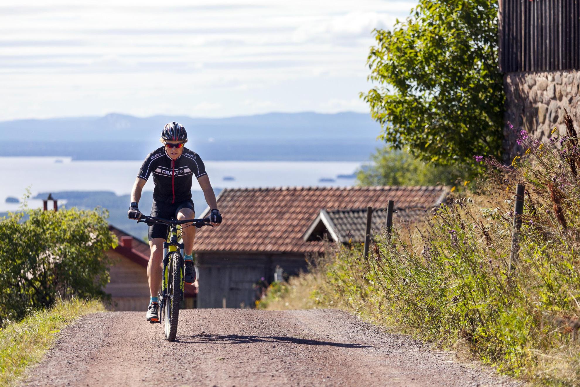 Een fietser in fietskleding die een heuvel op gaat, met houten huizen, heuvels en de zee op de achtergrond.