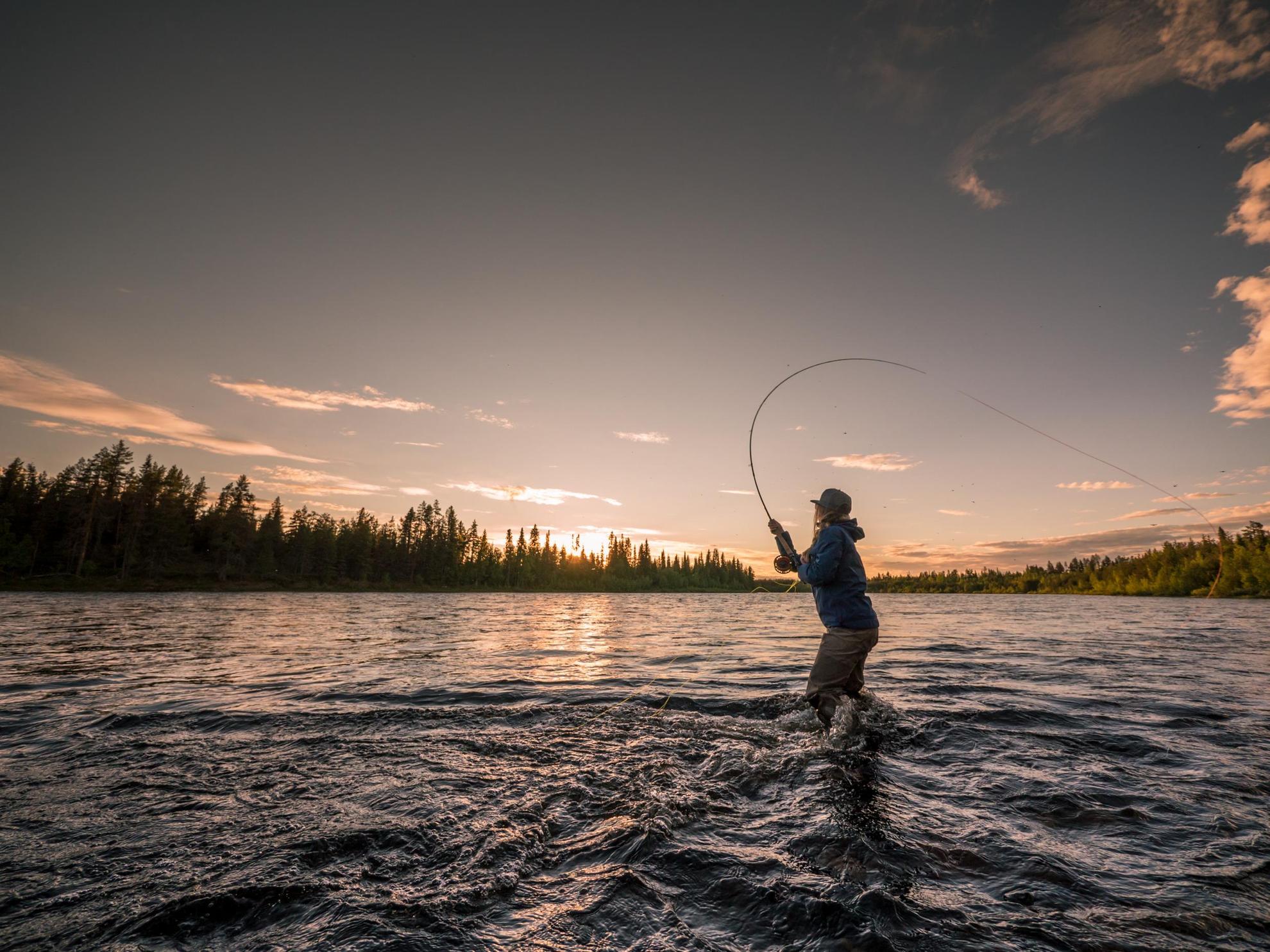 Een persoon is aan het vliegvissen en staat in een rivier in het noorden van Zweden terwijl de zon ondergaat.