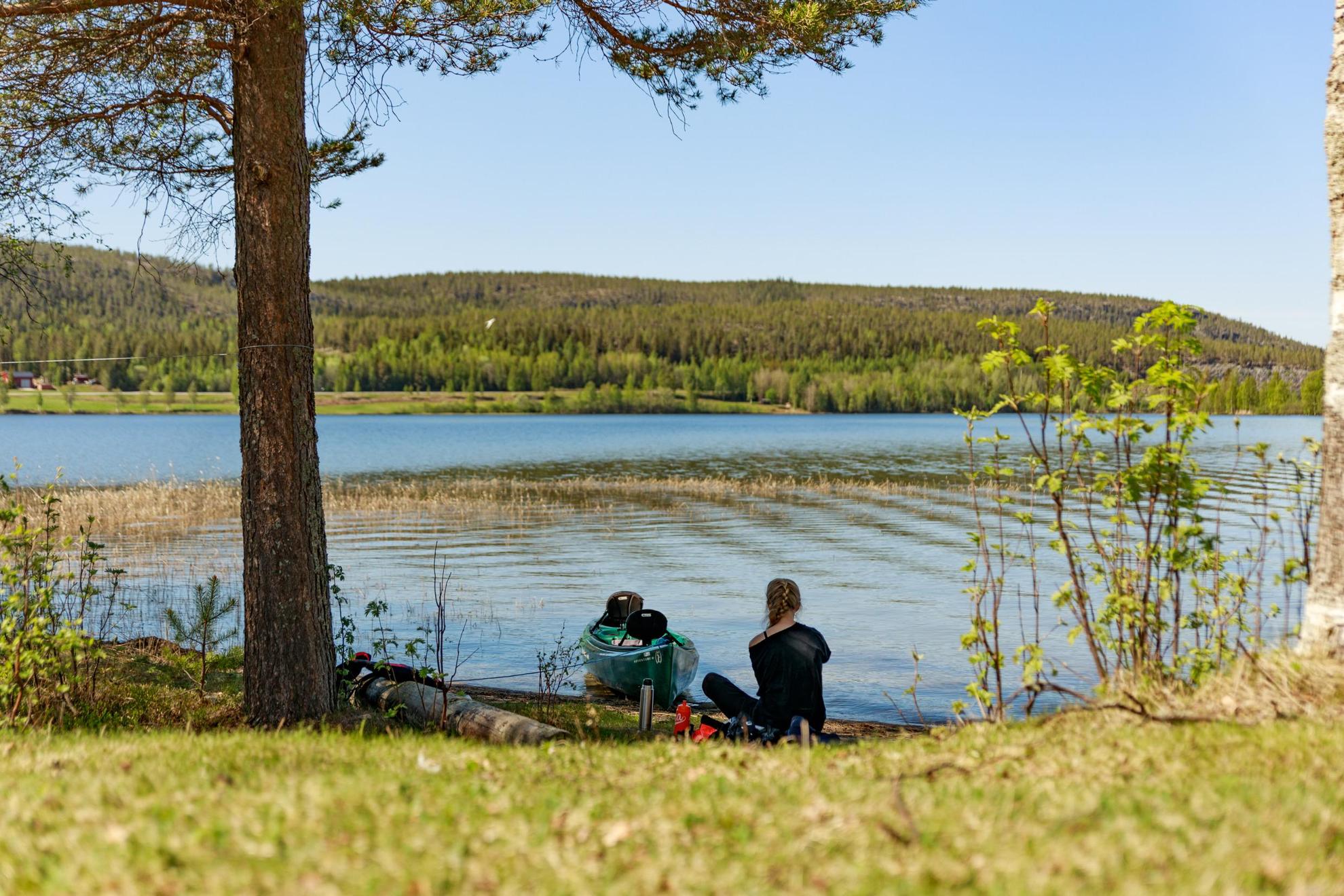 Een vrouw zit aan de oever bij een meer met haar kano afgemeerd aan de waterkant. Groot bos in de horizon aan de andere kant van het meer.