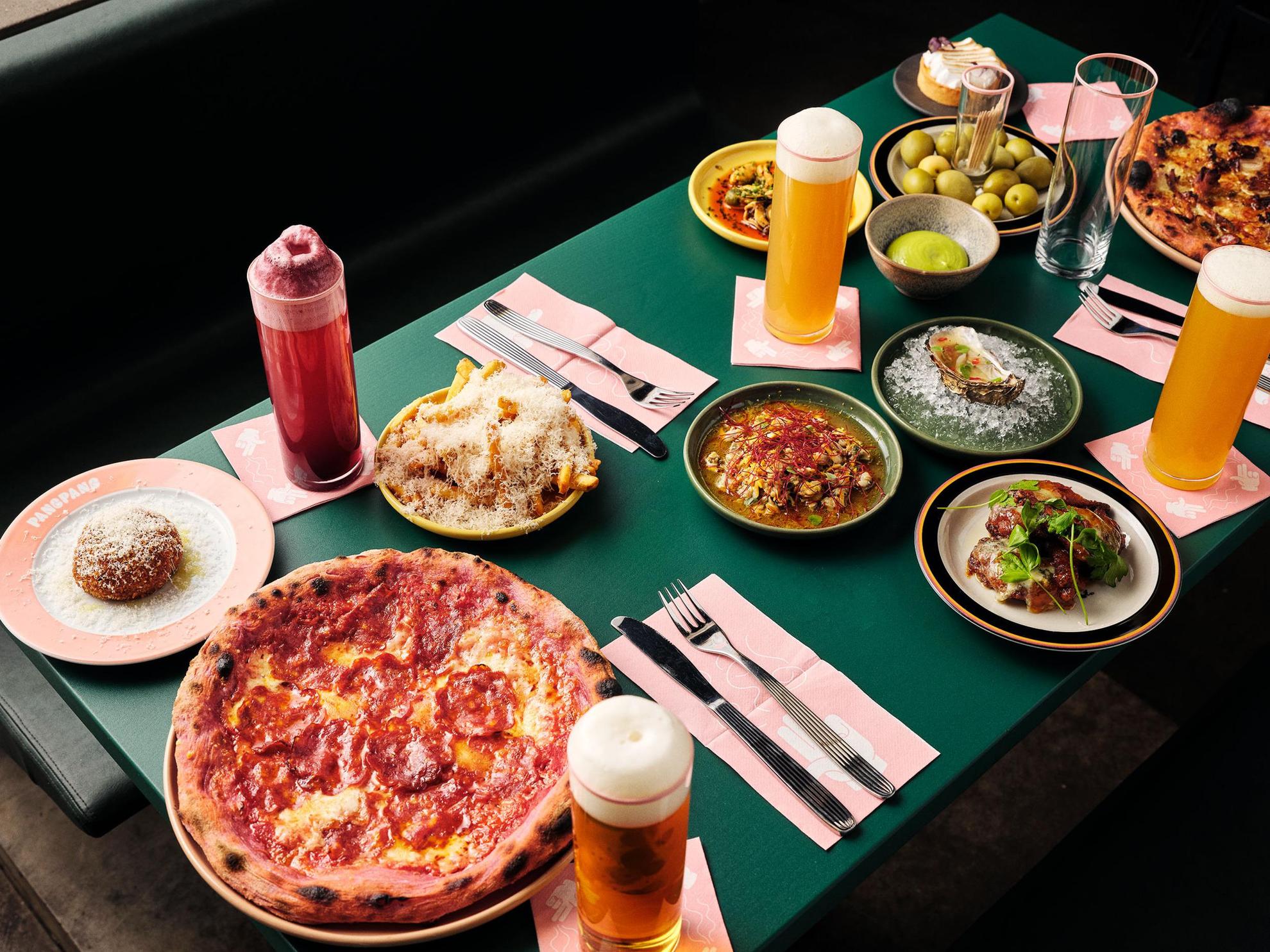 Een groene tafel met bestek, glazen gevuld met bier en borden met eten zoals pizza, frites, oesters en olijven.