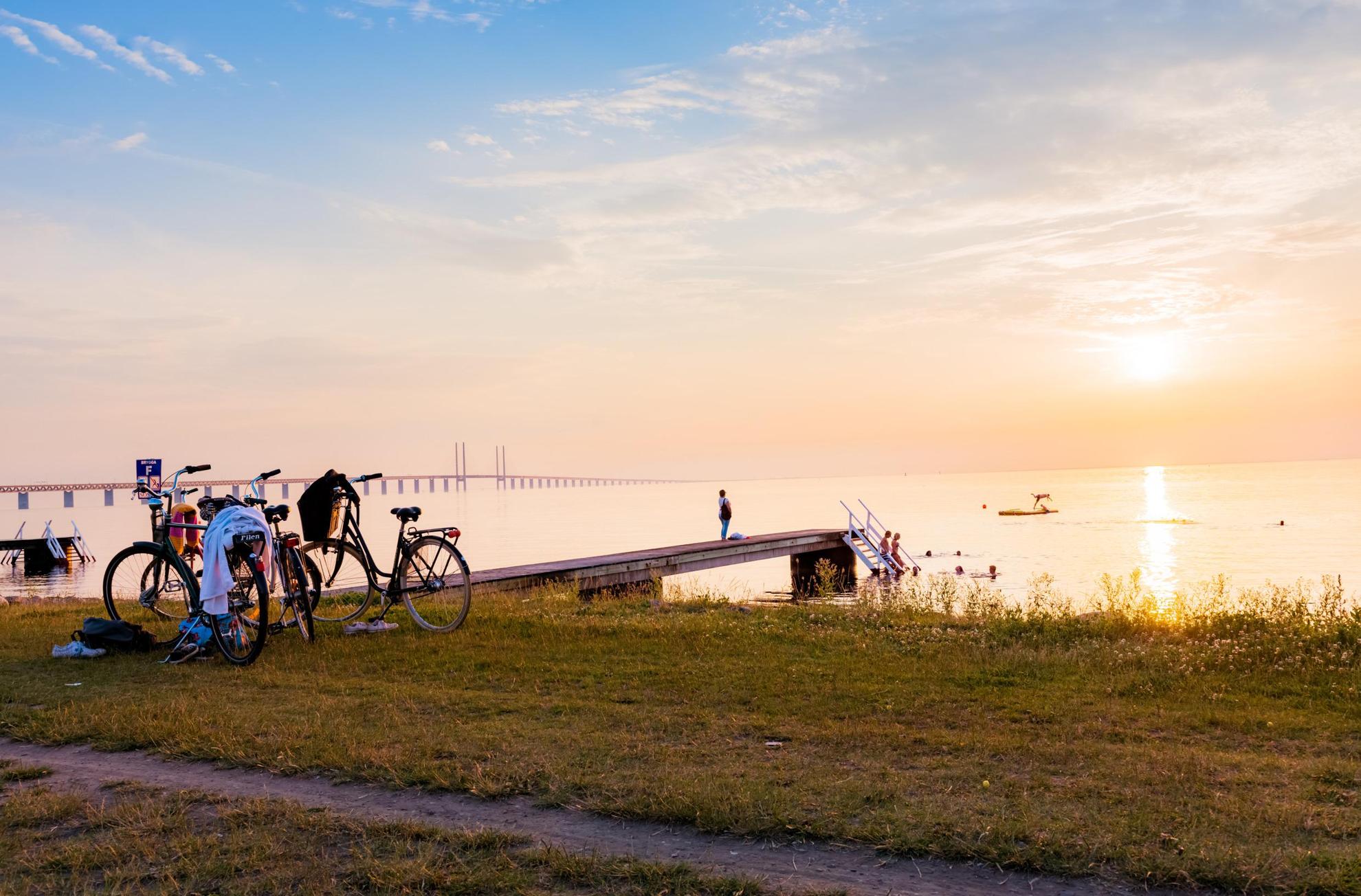 Zonsondergang boven een strand met een steiger. Op het gras boven het strand staan twee fietsen geparkeerd en meerdere mensen zwemmen in de zee. De Sontbrug op de achtergrond.