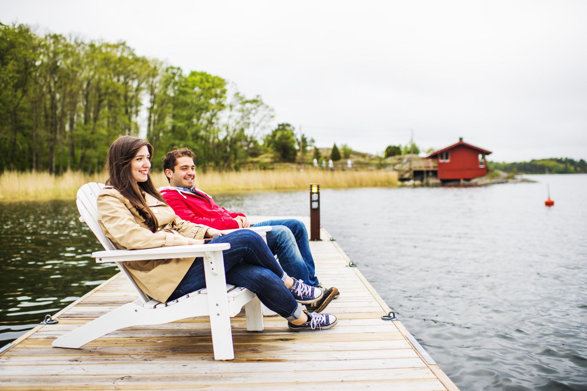 Twee mensen zitten op een houten stoel en kijken uit over het water. Achter hen zie je een rood huisje.